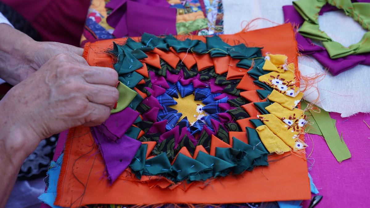 Mãos costurando retalhos coloridos de tecido em uma mesa de trabalho